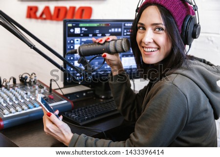 Beautiful happy young female radio host broadcasting in studio, using computer and headphones