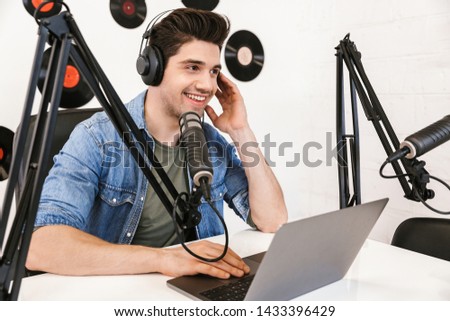 Happy young man radio host broadcasting through microphone in studio, using laptop computer