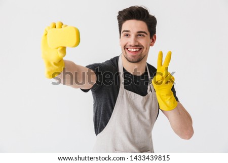 Handsome brunette houseman wearing apron standing isolated over white background, taking a selfie with a sponge