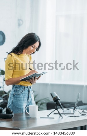 pretty radio host writing in notebook while standing near workplace in studio