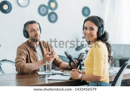 cheerful radio host looking at camera while sitting at workplace near colleague