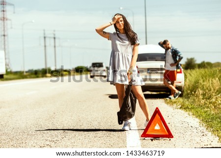 The young couple broke down the car while traveling on the way to rest. They are trying to stop other drivers and ask for help or hitchhike. Relationship, troubles on the road, vacation.