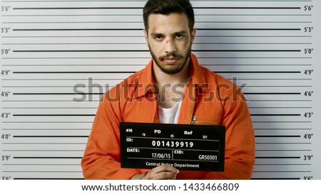 In a Police Station Arrested Man Getting Front-View Mug Shot. He's Wearing Prisoner Orange Jumpsuit and Holds Placard. Height Chart in the Background.