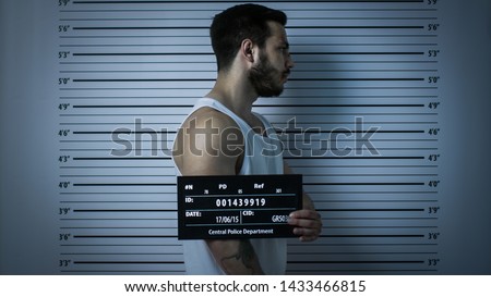In a Police Station Arrested Beaten Man Poses for Side View Mugshot. He Wears Singlet, is Heavily Bruised and Holds Placard. Height Chart in the Background. Shot with Dark Cold Lights, Vignette Filter Royalty-Free Stock Photo #1433466815