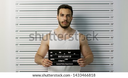 In a Police Station Arrested Beaten Man Poses for Front View Mugshot. He Wears Singlet, is Heavily Bruised and Holds Placard. Height Chart in the Background.