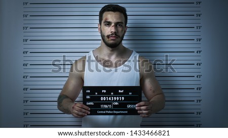 In a Police Station Arrested Beaten Man Poses for Front View Mugshot. He Wears Singlet, is Heavily Bruised and Holds Placard. Height Chart in the Background. Shot with Dark Cold Lights Vignette Filter
