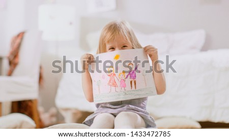 Cute Young Girl Sitting on Pillows and Shows Drawing of Her Family and Hides Behind It. Sunny Living Room.