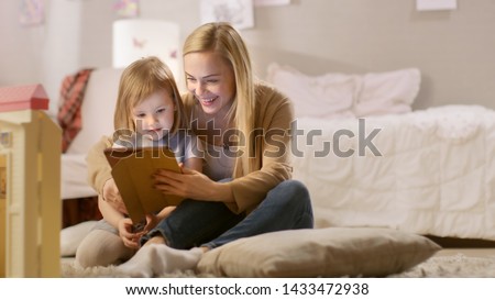 Beautiful Mother and Her Little Daughter Have Good Time Reading Children's Books on a Tablet Computer. Sunny Living Room.