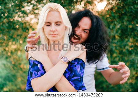Funny couple outdoor. Odd people. Crazy italian man frightening from behind praying woman with closed eyes.