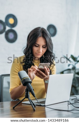 beautiful radio host using smartphone while sitting near microphone and laptop