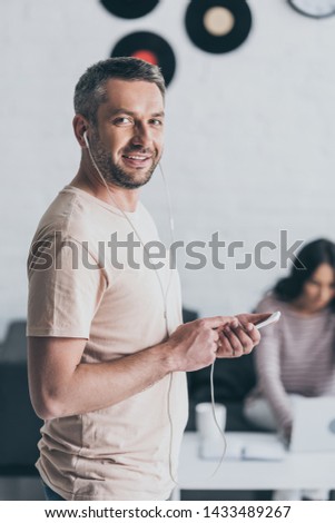 selective focus of cheeful man holding smartphone and listening music in earphones near young woman sitting at table with laptop