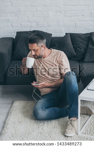 adult man sitting on floor, listening music in earphones, using smartphone and drinking coffee