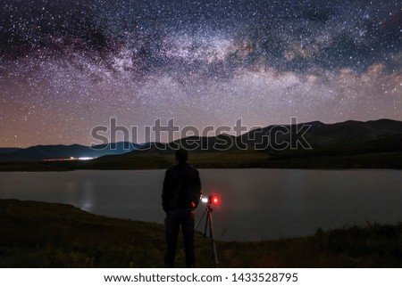 Man with camera stands on the shore of the lake in the starry night. Milky way galaxy background.