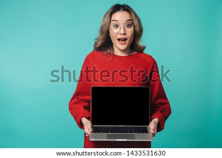 Portrait of a lovely excited young asian woman standing isolated over blue background, using blank screen laptop computer