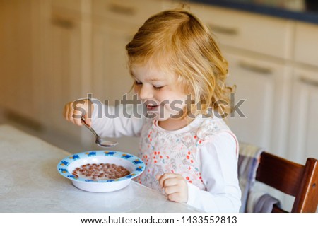 Adorable toddler girl eating healthy cereal with milk for breakfast. Cute happy baby child in colorful clothes sitting in kitchen and having fun with preparing oats, cereals. Indoors at home