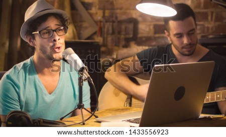 Two young man sing and play guitar while recording a song in a home studio in a garage.