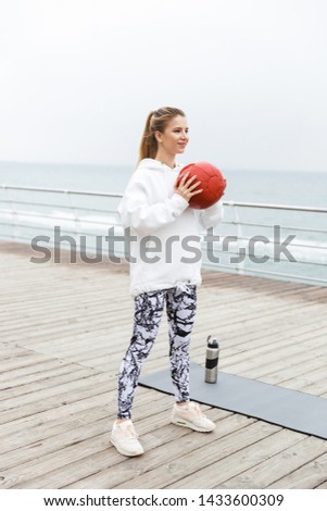 Image of attractive athletic woman 20s in white hoodie doing exercises with fitness boll while working out near seaside