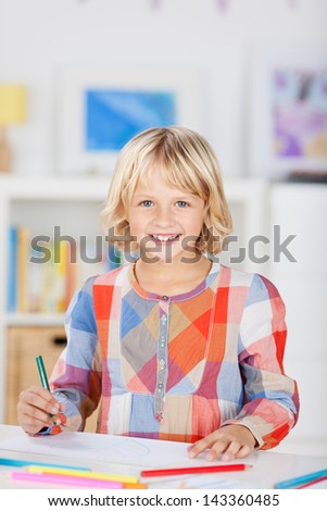 Small young blonde caucasian girl with a happy smile and wearing colorful clothes, draws a picture with color pencils at home