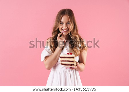Image of happy smiling woman wearing white dress looking at camera with her finger on her chin and holding piece of cake isolated over pink background
