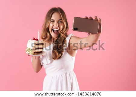 Image of young laughing woman taking selfie photo on cellphone and winking while holding piece of cake with candle isolated over pink background