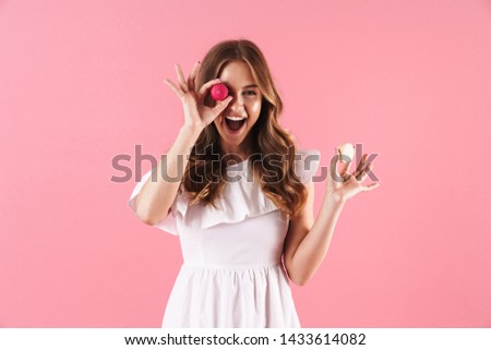 Image of happy joyful woman wearing white dress smiling at camera and holding macaroon biscuits isolated over pink background