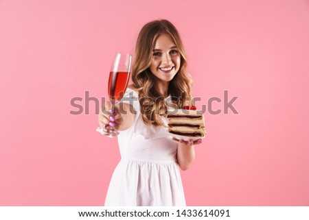 Image of cute smiling woman wearing white dress holding piece of cake and glass of wine isolated over pink background