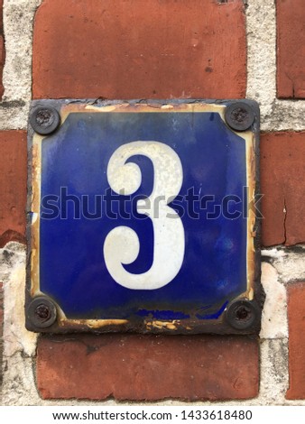 White house number 3 on a blue background, affixed to a red brick wall (early twentieth century). Garding, Schleswig-Holstein, Germany