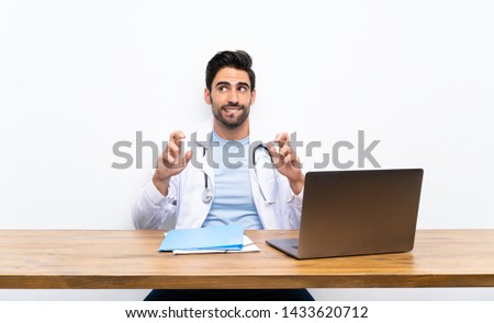 Young doctor man with his laptop over isolated wall with fingers crossing