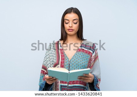 Young woman over isolated blue wall holding and reading a book