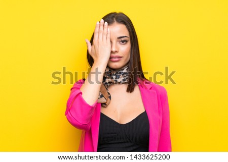 Young woman over isolated yellow background covering a eye by hand