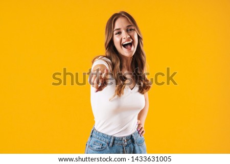 Image closeup of delighted caucasian woman wearing basic t-shirt smiling and pointing finger at camera isolated over yellow wall