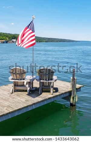 Two Adirondack Chairs on Wooden Pier with Waving American Flag and Background of Turquoise Lake Water, Tree Covered Hill and Blue Sky