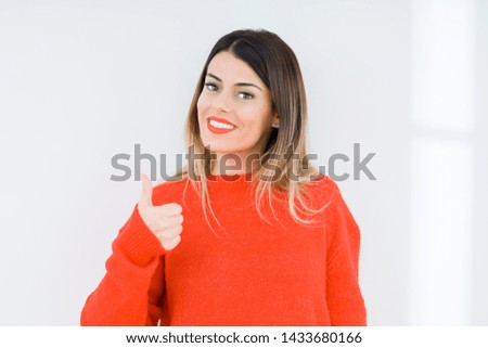 Young woman wearing casual red sweater over isolated background doing happy thumbs up gesture with hand. Approving expression looking at the camera showing success.