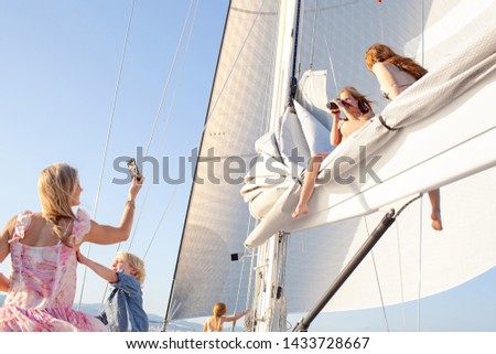 Family luxury sailing yacht summer holiday enjoying being at sea together, with girls up on mast taking pictures and mother using smartphone camera, travel fun outdoors. Leisure recreation lifestyle.