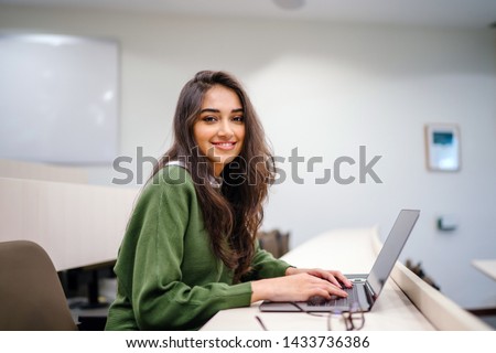 Portrait of a beautiful, young and intelligent-looking Indian Asian woman student wearing a white shirt and green tracker smiling as she works on her laptop in a university classroom.  Royalty-Free Stock Photo #1433736386