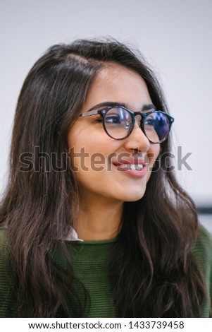 A close up head shot portrait of a preppy, young, beautiful, confident and attractive Indian Asian woman in a green sweater and spectacles against a white background. She is smiling happily. 