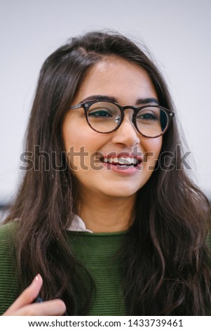 A close up head shot portrait of a preppy, young, beautiful, confident and attractive Indian Asian woman in a green sweater and spectacles against a white background. She is smiling happily. 