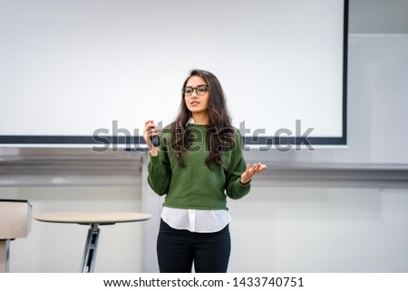 Portrait of a young, beautiful, attractive and intelligent Indian Asian woman wearing spectacles in a sweater giving a presentation in a lecture classroom. She is smiling as she is presenting. 