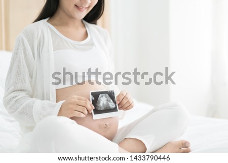 Pregnant woman holding the picture of the ultrasound on her belly in bed at bedroom. White background. Prenatal health care concept.