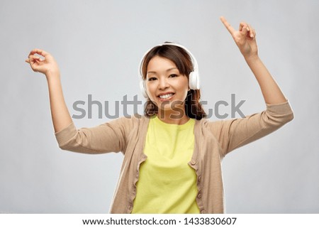 people, technology and audio equipment concept - happy young asian woman in headphones listening to music and dancing over grey background