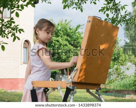 A little girl and her great-grandmother play together in the garden and draw. Easel, canvas, brushes and paints. Recreation and summer games in the open air. Granny.