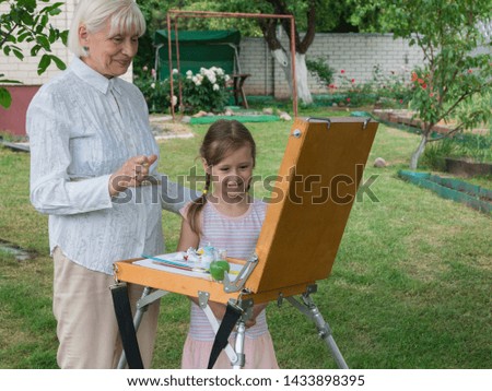 A little girl and her great-grandmother play together in the garden and draw. Easel, canvas, brushes and paints. Recreation and summer games in the open air. Granny.