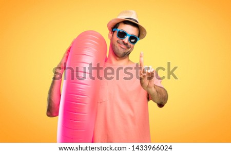Man with hat and sunglasses on his summer vacation showing and lifting a finger in sign of the best on orange background