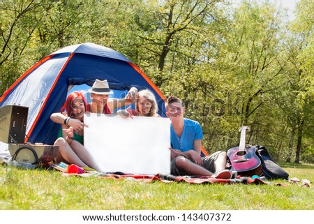 Four teenagers in colored shirts on the camping with a white board which can be used for your advertisements