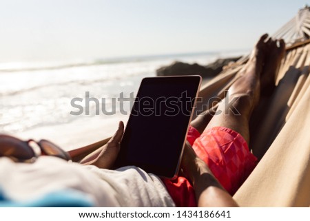 Low section of African-american man using digital tablet while relaxing in a hammock on the beach Royalty-Free Stock Photo #1434186641