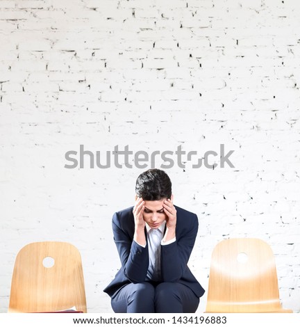Young businesswoman with headache sitting against white brick wall at office lobby