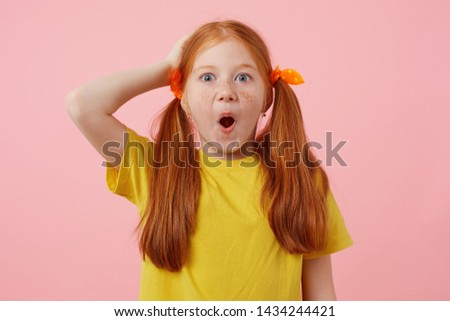 Portrait of happy amazed petite freckles red-haired girl with two tails, wears in yellow t-shirt, stands over pink background with wide open mouth.