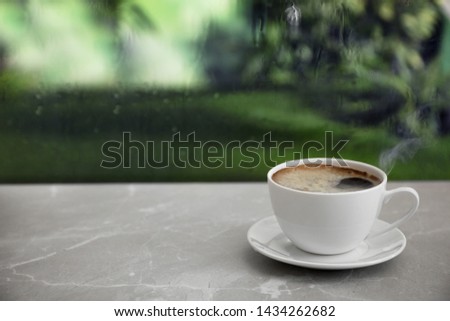 Cup of hot drink on stone windowsill against glass with rain drops, space for text