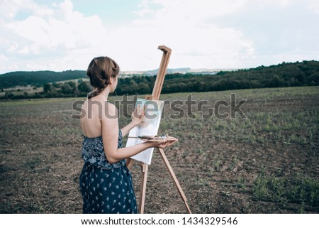 Beautiful girl in vintage dress creating a painting outdoors, Young artist near easel and canvas in the field, Gorgeous womain in blue dress painting near easel, braided hair