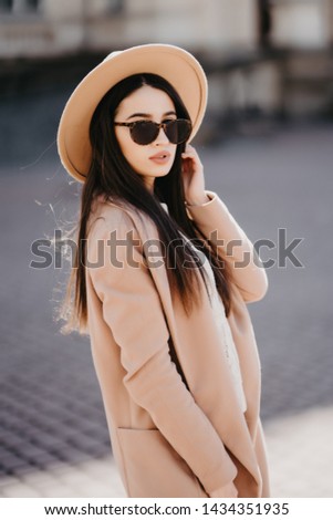 Beautiful young hipster girl with sunglasses in a coat with a bag walking in the spring city at sunset